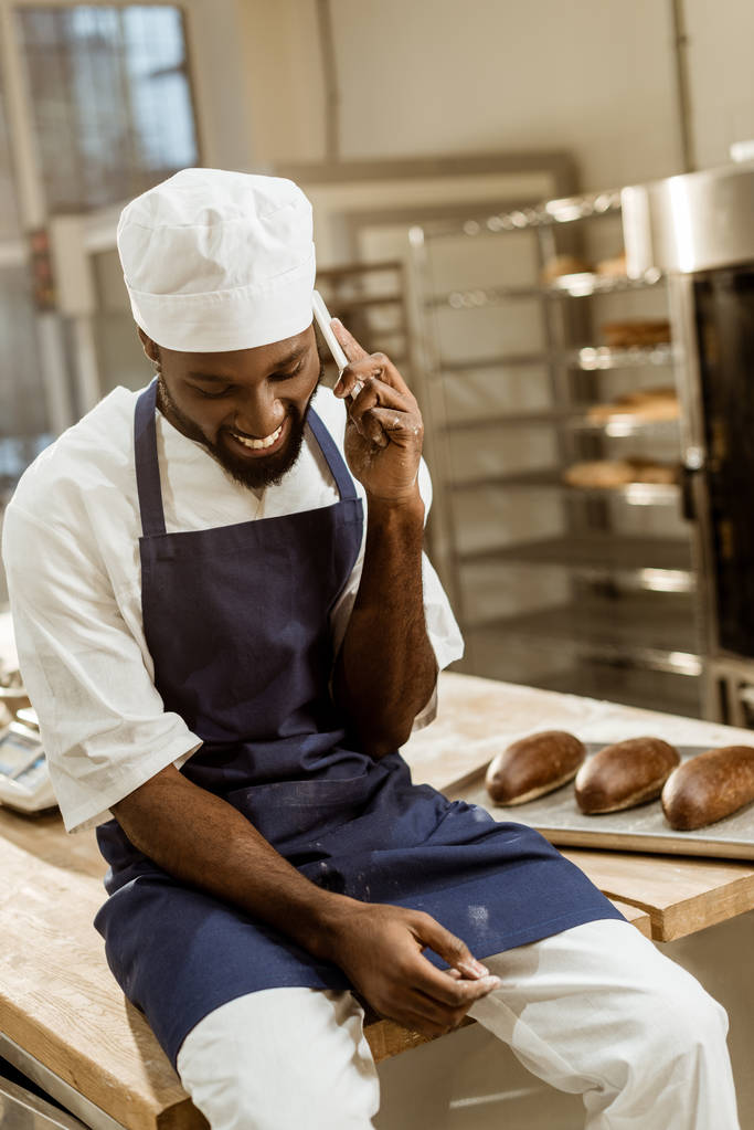 mladí africké americké baker s rukama obalený v mouce, rozhovor po telefonu a zároveň sedí na pracovním stole - Fotografie, Obrázek