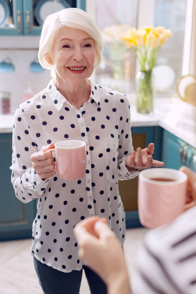 Iäkäs nainen juo kahvia keittiössä ja puhuu
 - Valokuva, kuva
