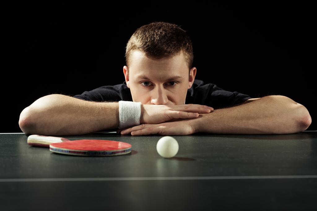 portrait of thoughtful tennis player at tennis table with ball and racket isolated on black - Photo, Image