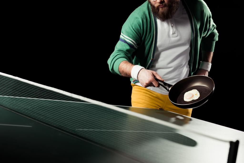 partial view of tennis player holding frying pan with egg in hand at tennis table isolated on black - Photo, Image