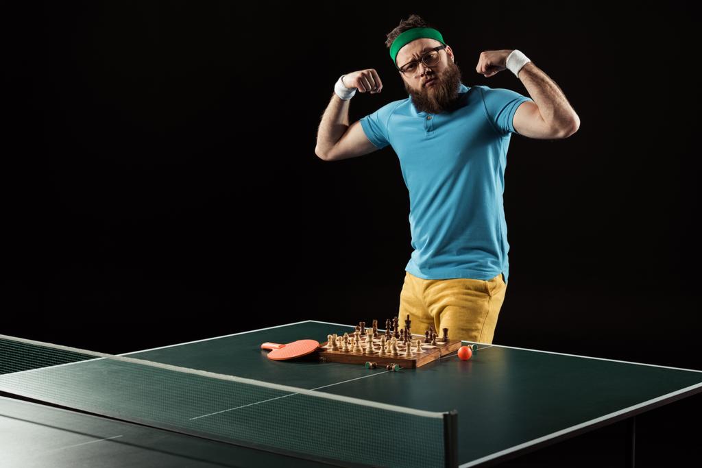 tennis player showing muscles while standing at tennis table with chess board isolated on black - Photo, Image