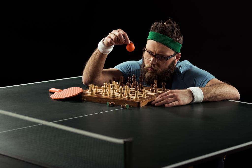 desportista barbudo jogando bola de tênis no tabuleiro de xadrez na mesa de tênis isolado em preto
 - Foto, Imagem