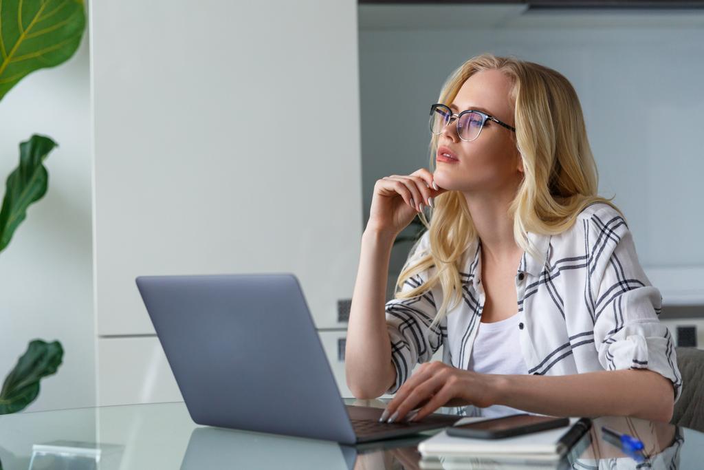 mooie peinzende blonde vrouw laptop gebruikt terwijl u thuis werkt - Foto, afbeelding