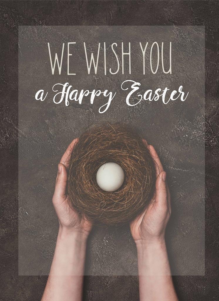 обрезанный вид человека, держащего гнездо с белым яйцом, и Мы желаем вам счастливой Пасхи надписи
 - Фото, изображение