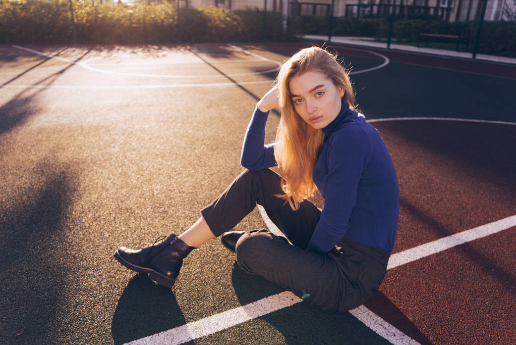 стильная красивая девушка модель блондинка в голубом свитере сидит на спортивной площадке на земле и позирует
 - Фото, изображение