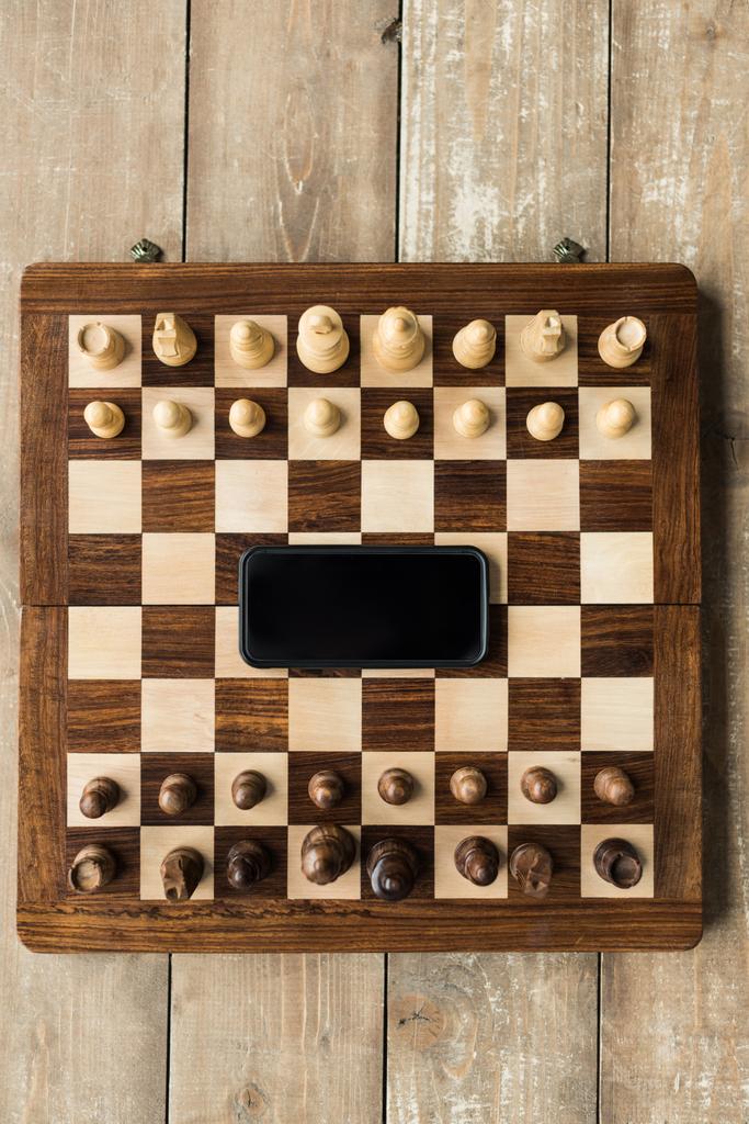 Vista superior del tablero de ajedrez con smartphone y piezas de ajedrez sobre superficie de madera
 - Foto, imagen