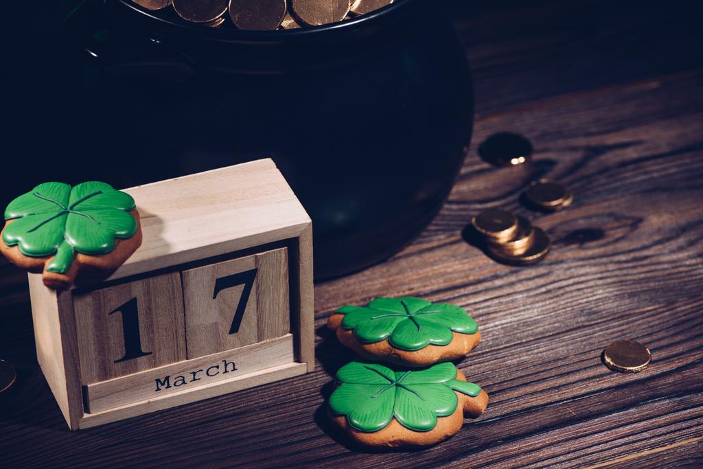 Крупный план календаря, печенье в форме трилистника и горшок с золотыми монетами на деревянном столе
   - Фото, изображение