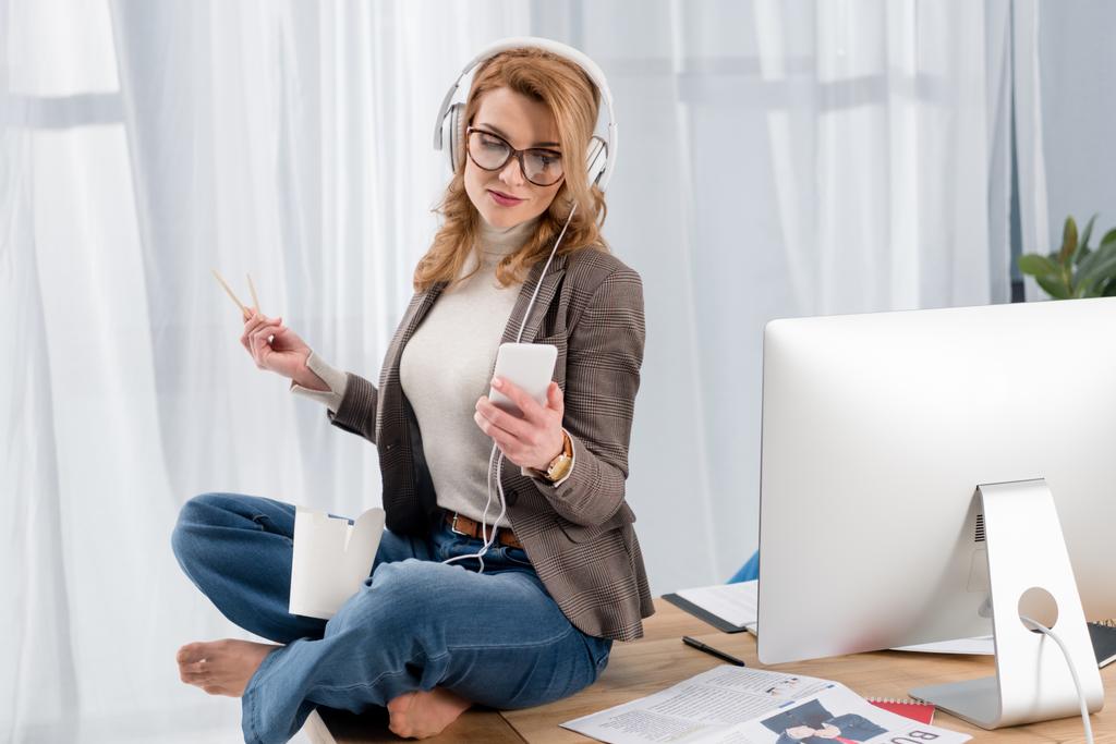 Geschäftsfrau mit Kopfhörer und Smartphone hört Musik auf Tisch im Büro - Foto, Bild