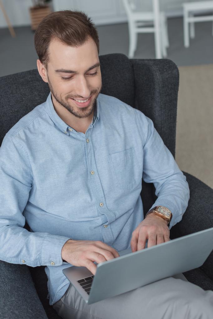 Portret van glimlachende man met behulp van de laptop tijdens het rusten in leunstoel - Foto, afbeelding