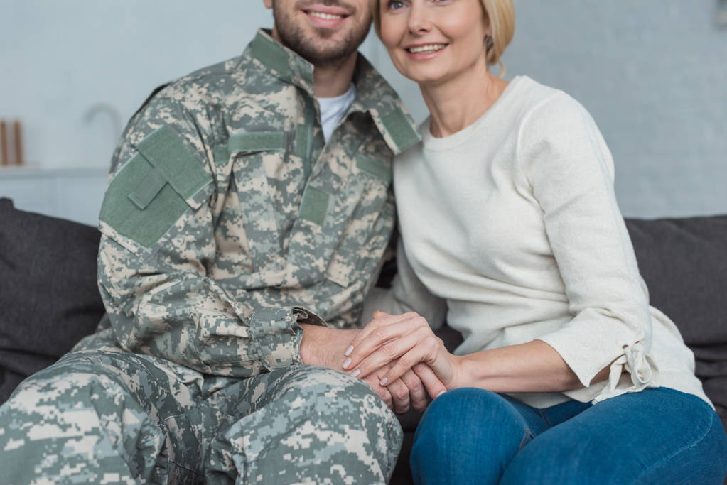 partial view of smiling mother and grown son in military uniform holding hands on sofa at home - Photo, Image