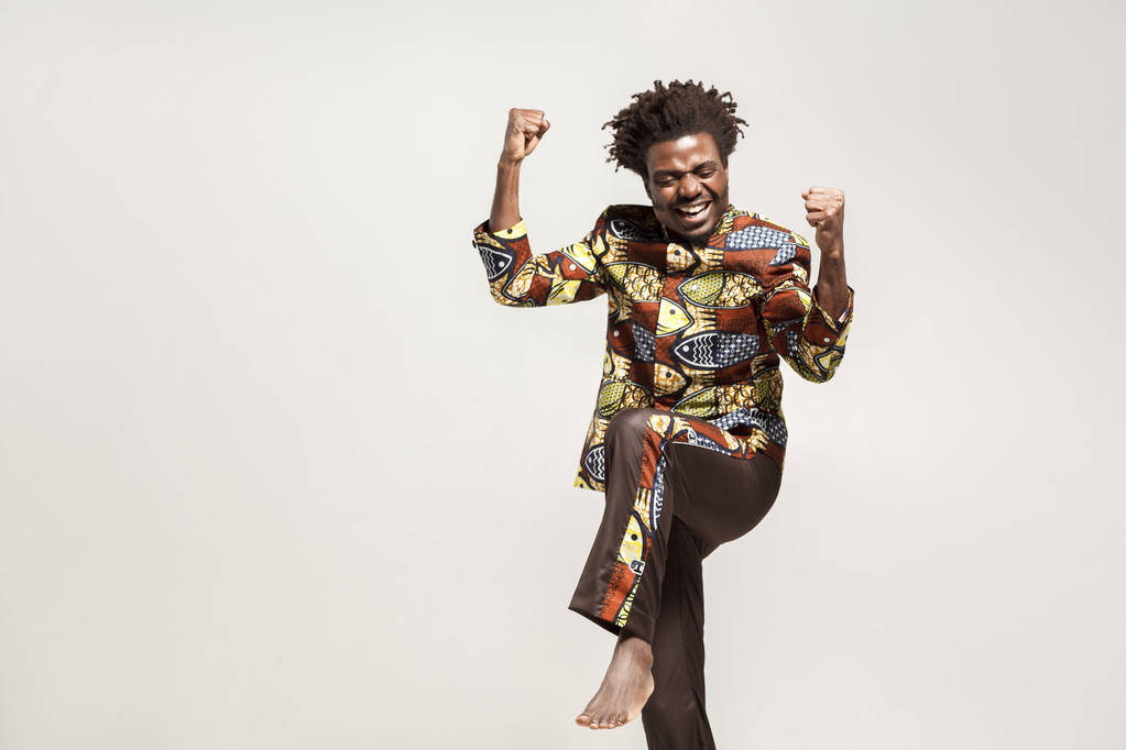onnellinen optimistinen afrikkalainen amerikkalainen mies perinteisissä vaatteissa nosti kädet riemuiten voitto harmaalla pohjalla
 - Valokuva, kuva