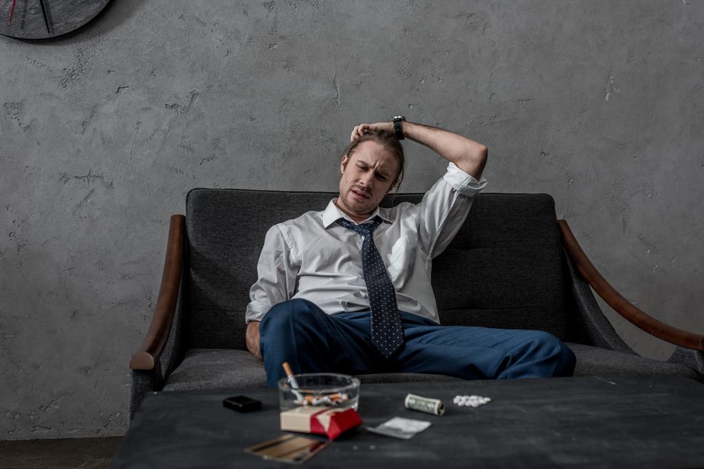 κατάθλιψη επιχειρηματίας με την τοξικομανία, που κάθεται στον καναπέ μπροστά από τον πίνακα με τα ναρκωτικά - Φωτογραφία, εικόνα