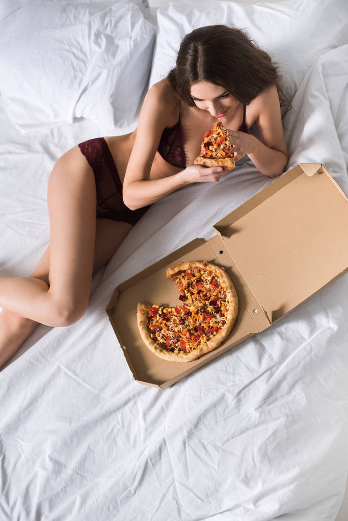 Hochwinkelaufnahme der verführerischen Frau, die in Spitzenunterwäsche auf dem Bett liegt und Pizza isst - Foto, Bild