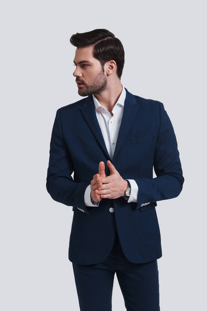 joven guapo en traje mirando hacia otro lado mientras está de pie sobre fondo gris con las manos apretadas
 - Foto, Imagen