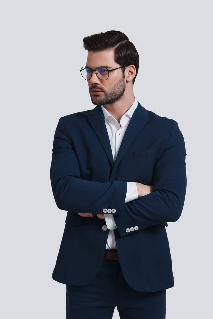 mladý a úspěšný, pohledný muž v obleku drží paže překřížené a vypadající při postavení proti šedé pozadí - Fotografie, Obrázek
