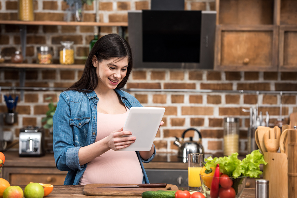 sorrindo jovem grávida usando tablet digital enquanto cozinha na cozinha
 - Foto, Imagem
