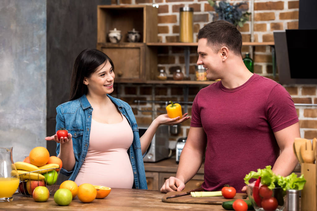 Szczęśliwa Młoda para w ciąży uśmiechający się wzajemnie podczas gotowania razem w kuchni  - Zdjęcie, obraz