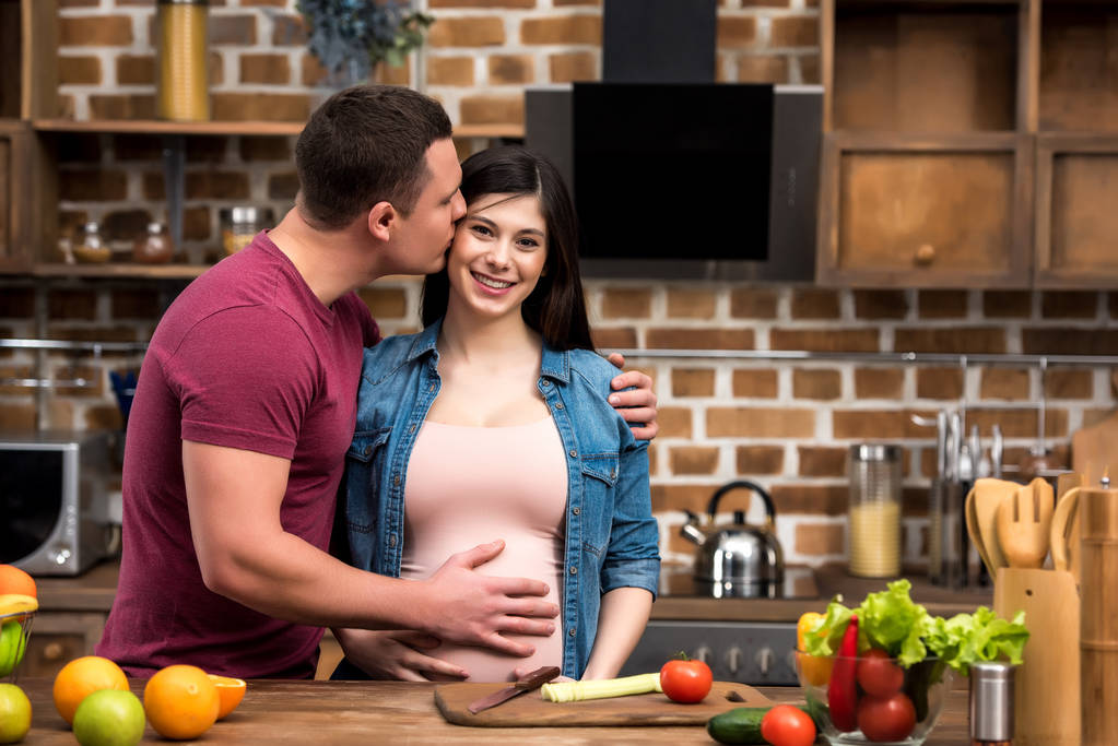 jovem abraçando e beijando feliz esposa grávida na cozinha
 - Foto, Imagem