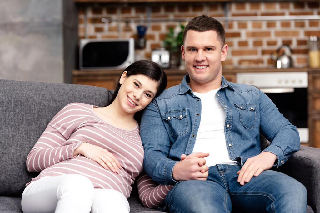 счастливая молодая беременная пара, сидящая вместе на диване и улыбающаяся в камеру
 - Фото, изображение