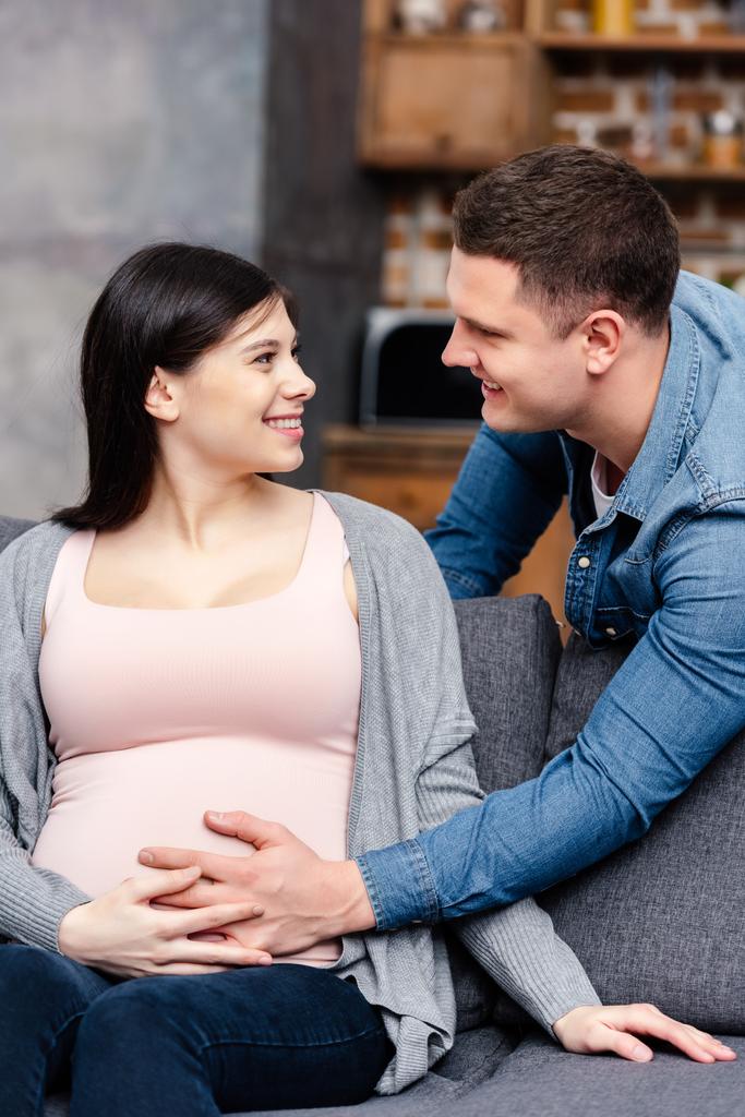 gelukkige jonge zwangere paar aanraken van de buik en glimlachen elkaar thuis - Foto, afbeelding