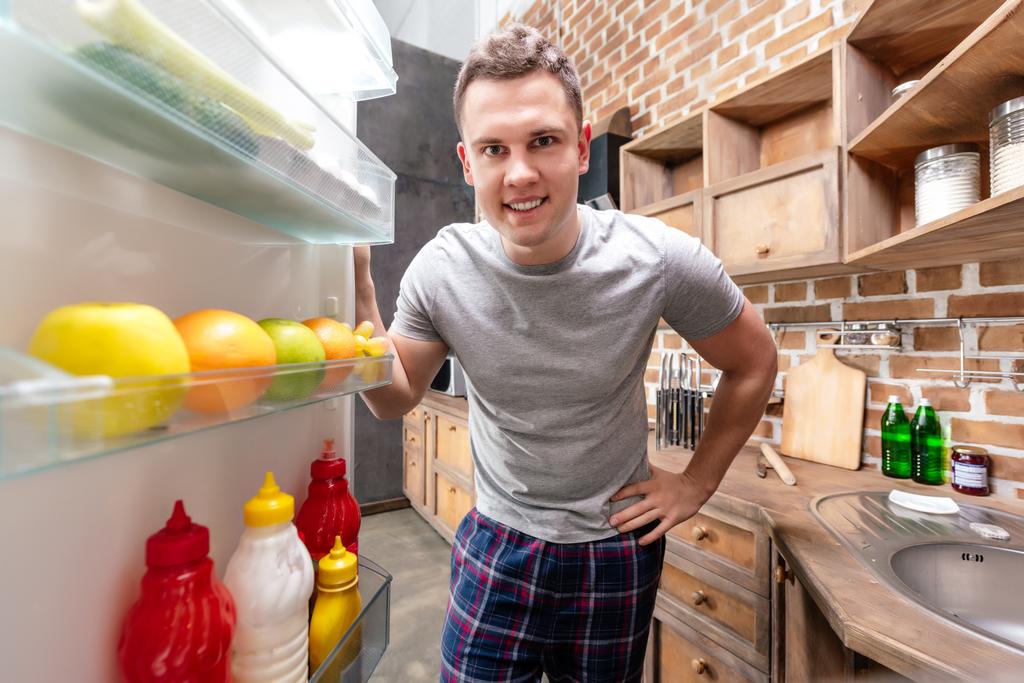 guapo joven sonriente en pijama mirando en el refrigerador
 - Foto, imagen