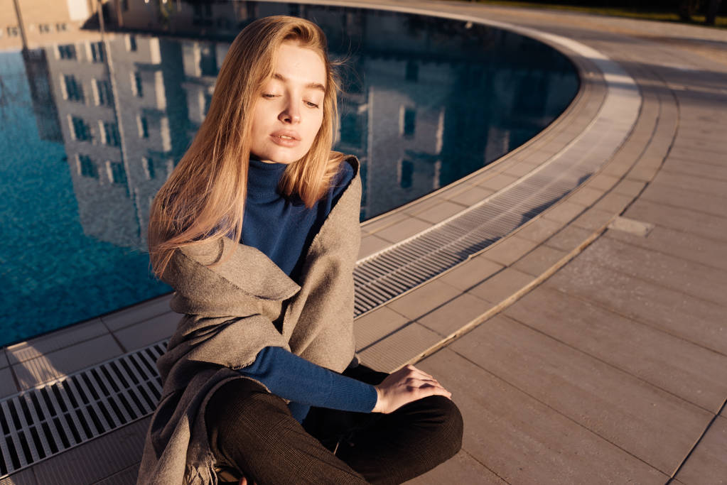 Dziewczyna w niebieskim swetrze siedzi przy basenie, cieszy się słońcem - Zdjęcie, obraz
