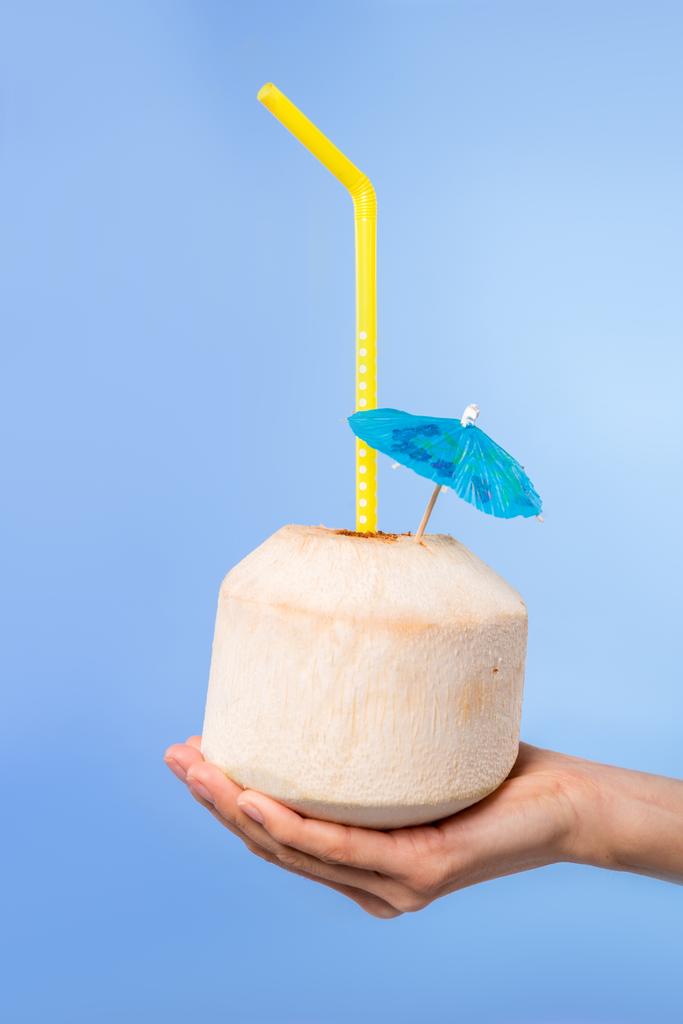 abgeschnittene Ansicht der weiblichen Hand mit Sommercocktail in frischer Kokosnuss mit Regenschirm und Stroh, isoliert auf blau - Foto, Bild