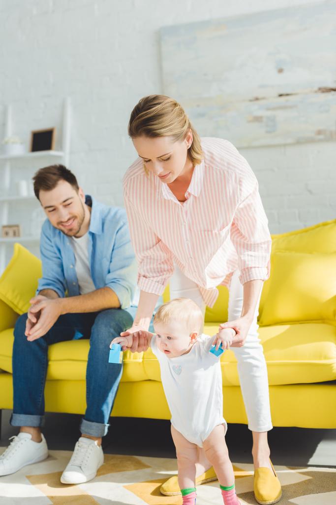 Mother teaching infant daughter to walk and father sitting on couch in modern room - Photo, Image