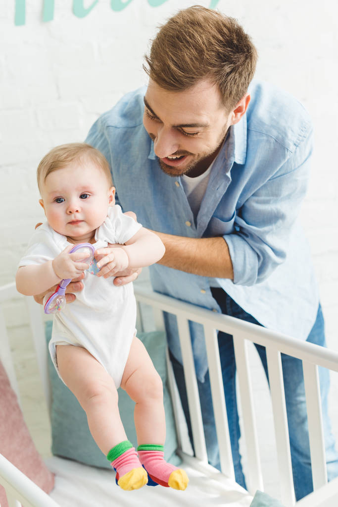 Happy father holding infant daughter with toy in crib - Photo, Image