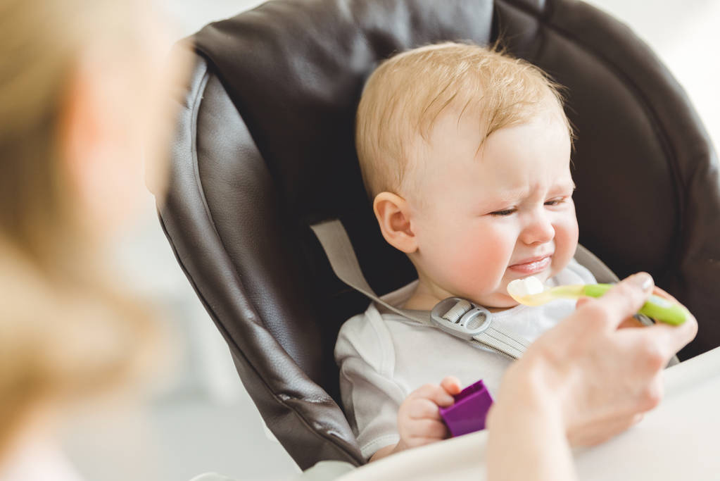 Hija lactante en silla de bebé rechazando la comida que la madre le da
  - Foto, Imagen