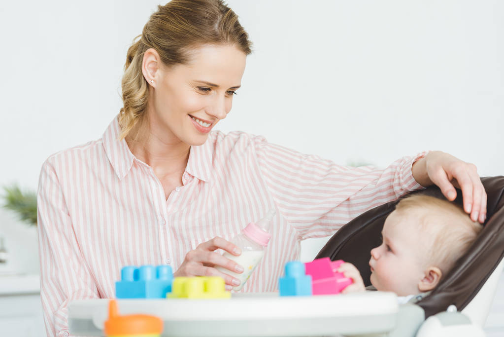 Moeder met voedingsfles en dochtertje zit in kinderstoel met kunststof blokken - Foto, afbeelding