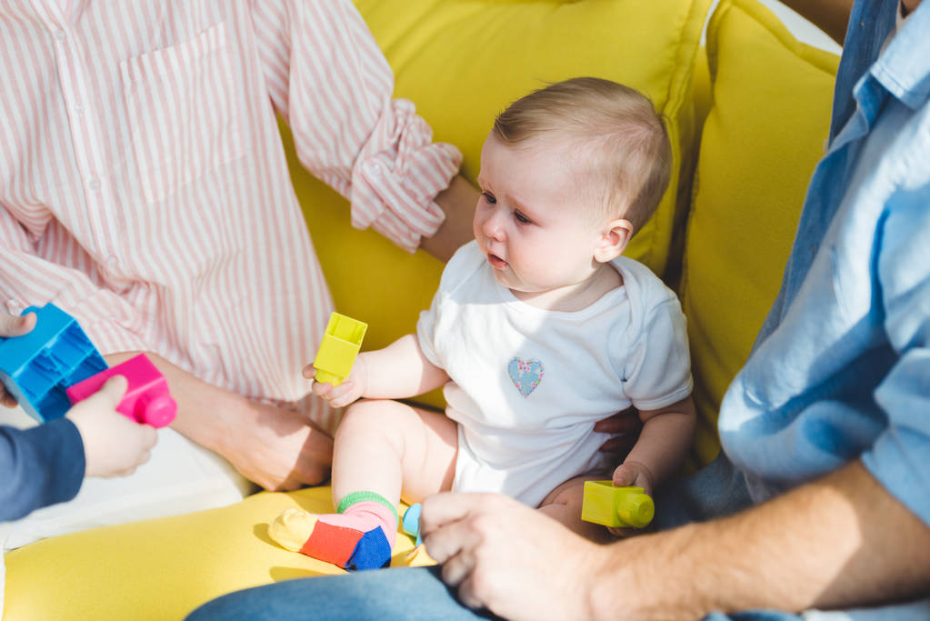 Обрізаний вид батьків, що сидять на дивані з дочкою-немовлям, що тримає пластикові блоки
 - Фото, зображення