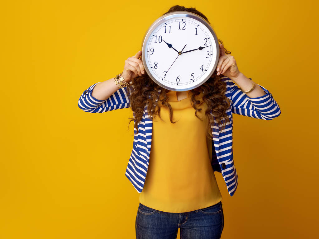 young woman in striped jacket on yellow background holding clock in the front of face - Photo, Image