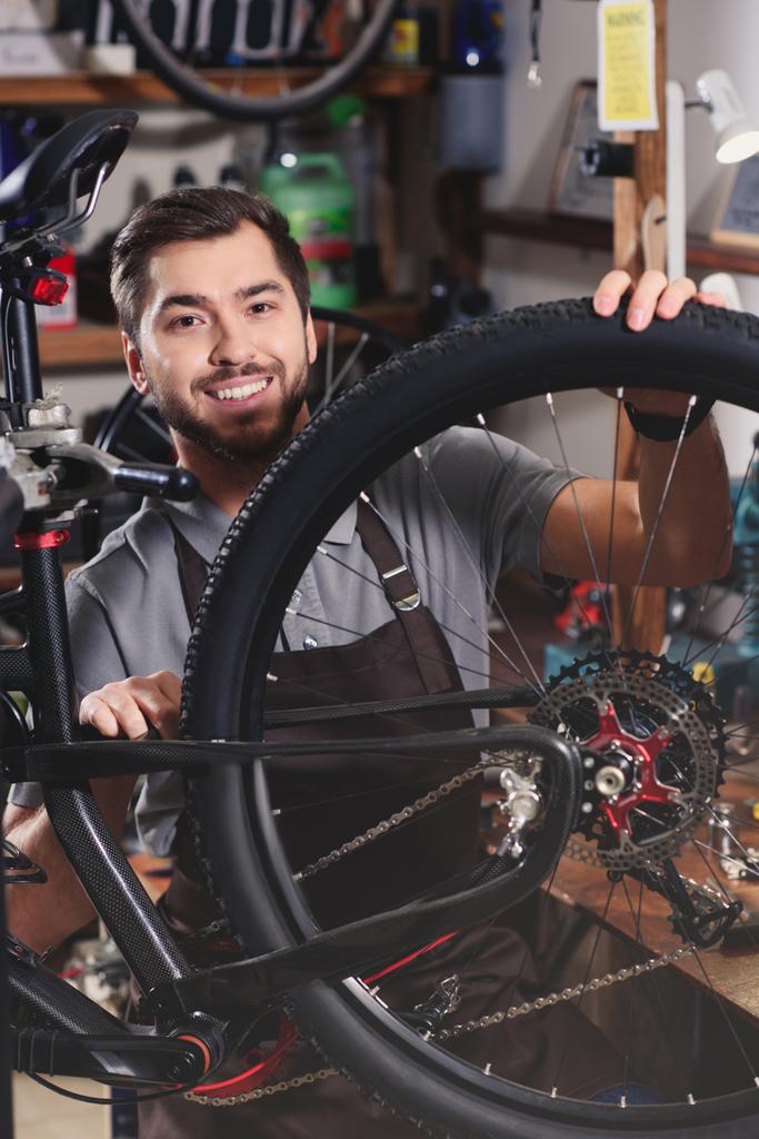 joven guapo en delantal fijación de bicicleta y sonriendo a la cámara en el taller
 - Foto, Imagen