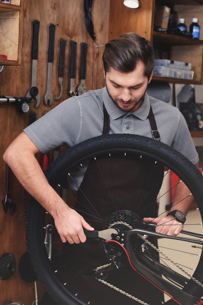 молодой человек в фартуке фиксирует велосипедное колесо и цепь с гаечным ключом в мастерской
 - Фото, изображение