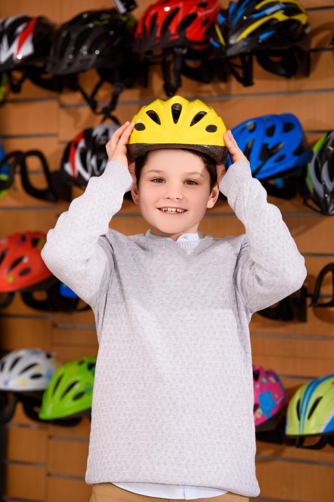 ładny mały chłopiec, stawiając na kask rowerowy i uśmiecha się do kamery w sklepie rowerowym  - Zdjęcie, obraz