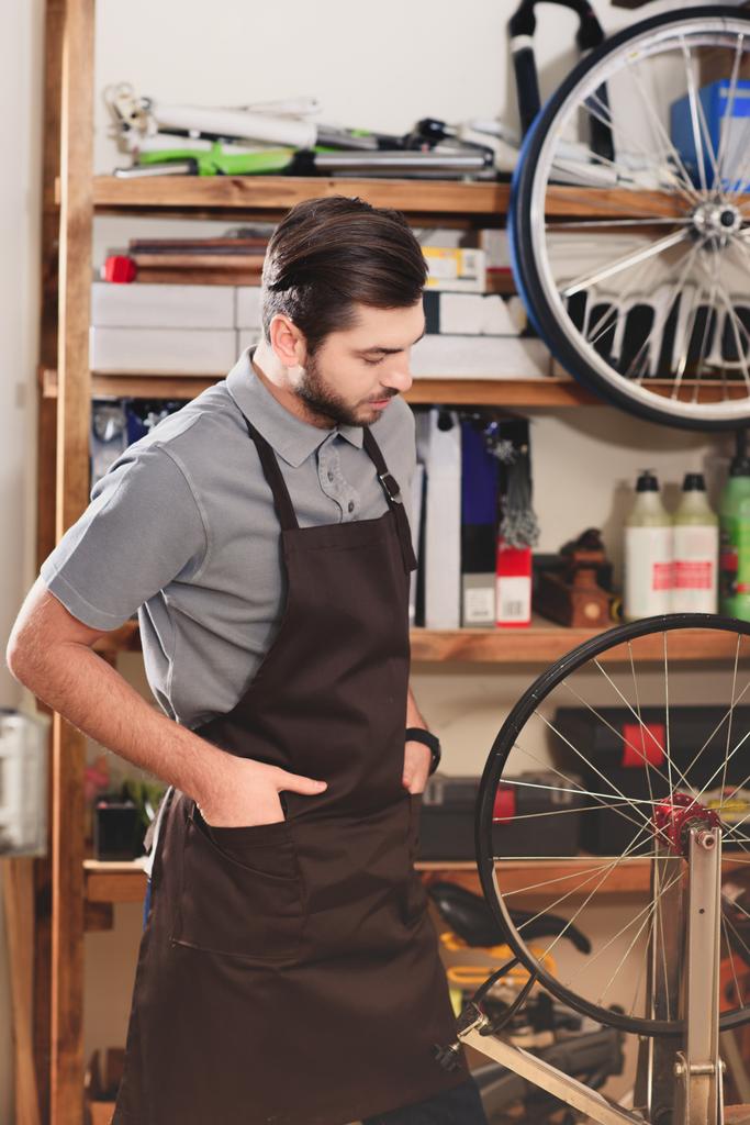 joven trabajador confiado en delantal mirando rueda de bicicleta en taller
 - Foto, imagen
