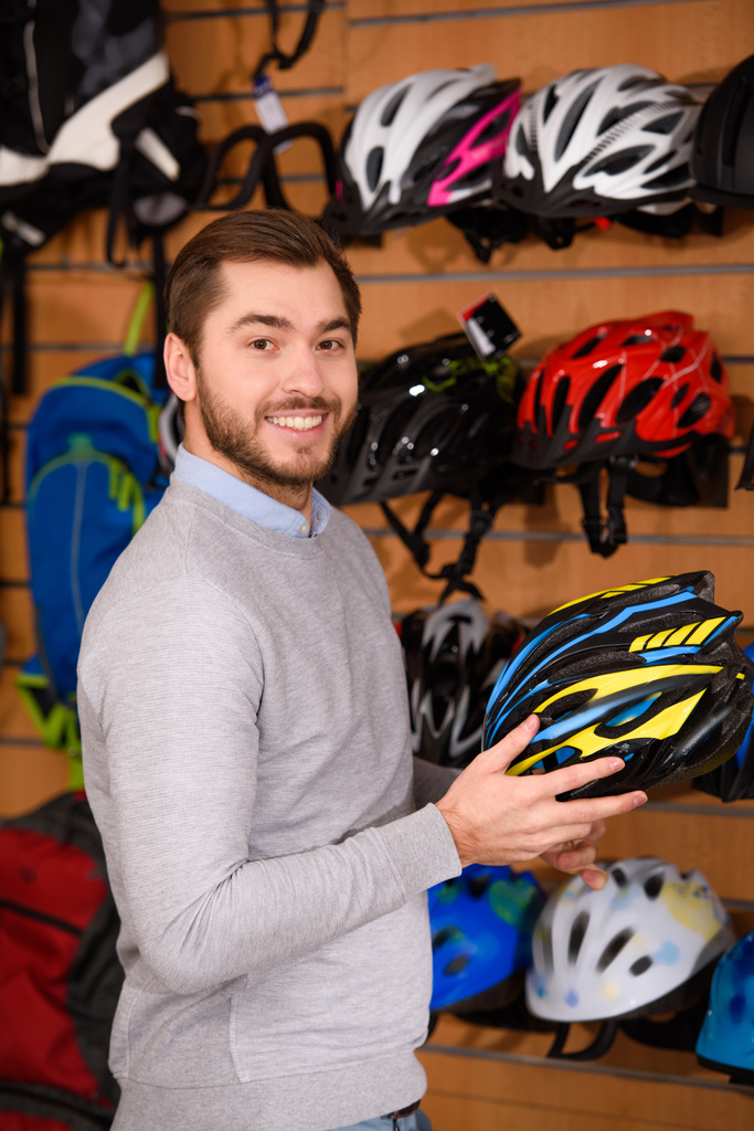 όμορφος νεαρός άνδρας κρατά κράνος ποδηλάτου και να χαμογελά στη φωτογραφική μηχανή στο κατάστημα ποδηλάτων - Φωτογραφία, εικόνα