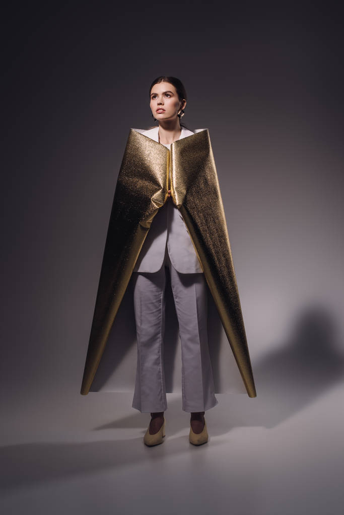 κομψή γυναίκα με λευκό κοστούμι που καλύπτονται σε χρυσό αναδίπλωσης χαρτιού θέτοντας σε σκούρο φόντο - Φωτογραφία, εικόνα