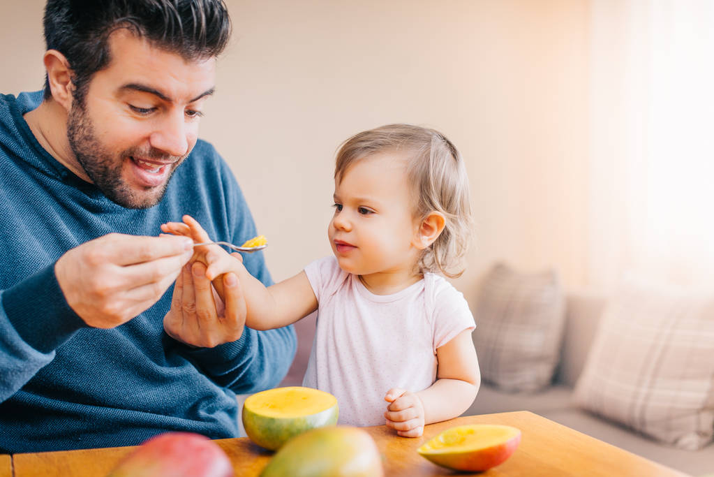 close-up of young happy father feeding cute little girl with fresh mango at home  - Photo, Image