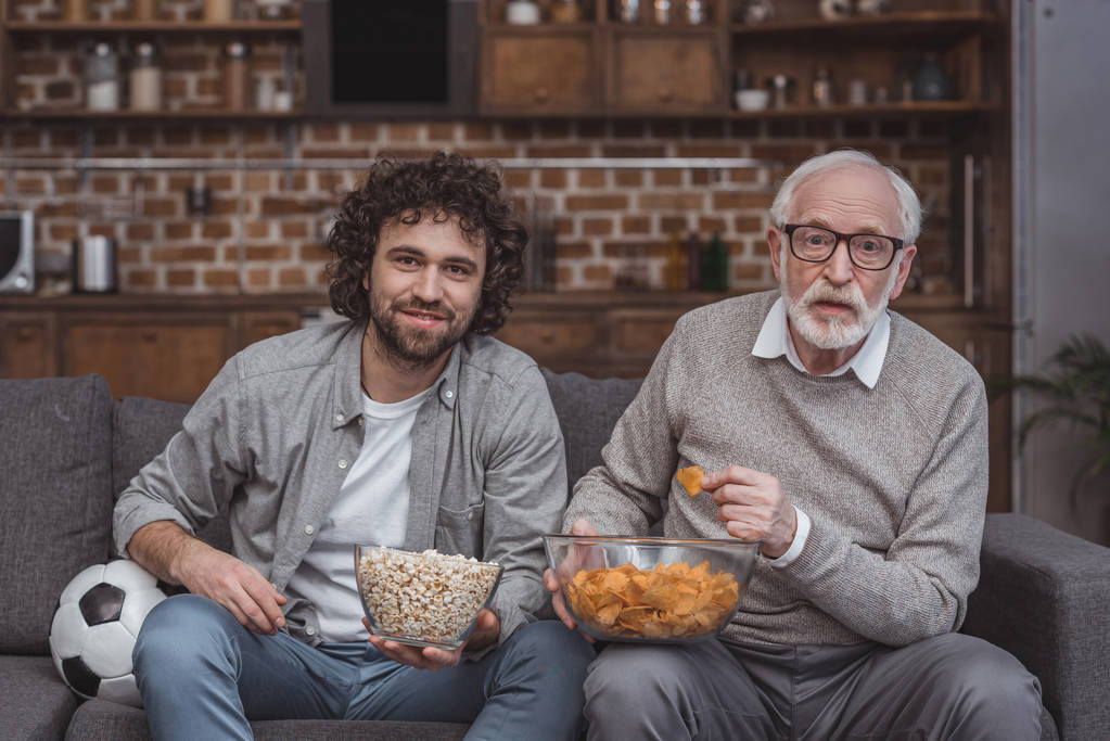 взрослый сын и старший отец смотрят футбол с попкорном и чипсами дома
 - Фото, изображение