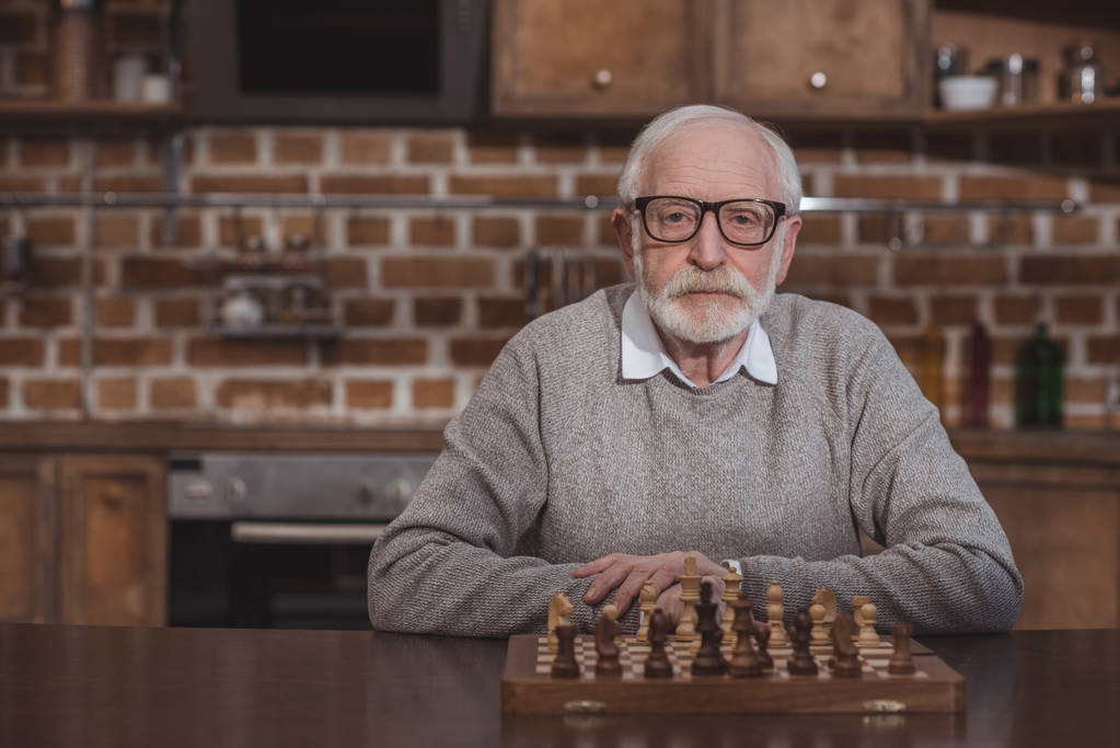 guapo hombre de pelo gris sentado en la mesa con tablero de ajedrez y mirando a la cámara
 - Foto, imagen