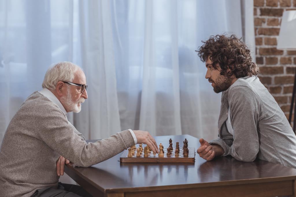 side view of adult son and senior father playing chess at home - Photo, Image