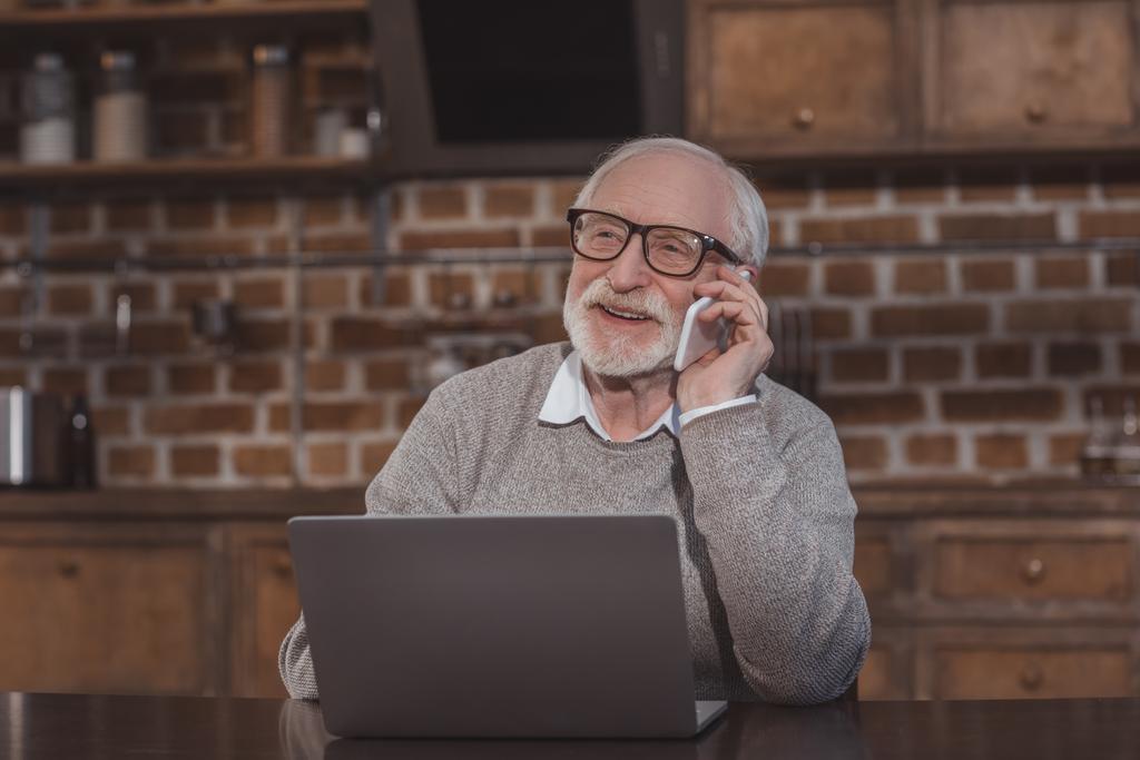 sorrindo bonito homem de cabelo cinza falando por smartphone à mesa com laptop
 - Foto, Imagem