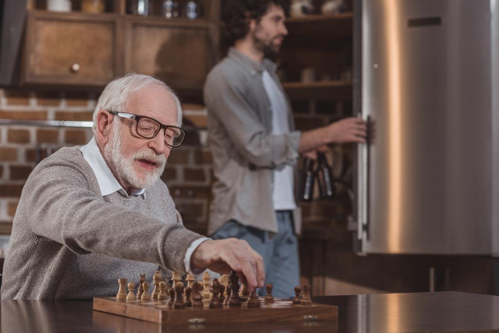 filho adulto olhando na geladeira e pai sênior jogando xadrez em casa
 - Foto, Imagem
