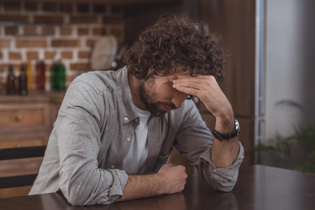 tired man sitting at table in kitchen and touching head - Photo, Image