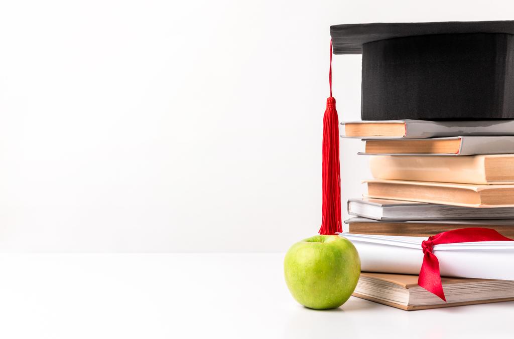 Academic hat on pile of books with diploma and apple isolated on white - Photo, Image