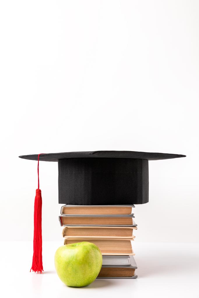 Яблуко біля купи книг з академічною шапочкою зверху ізольовано на білому
 - Фото, зображення