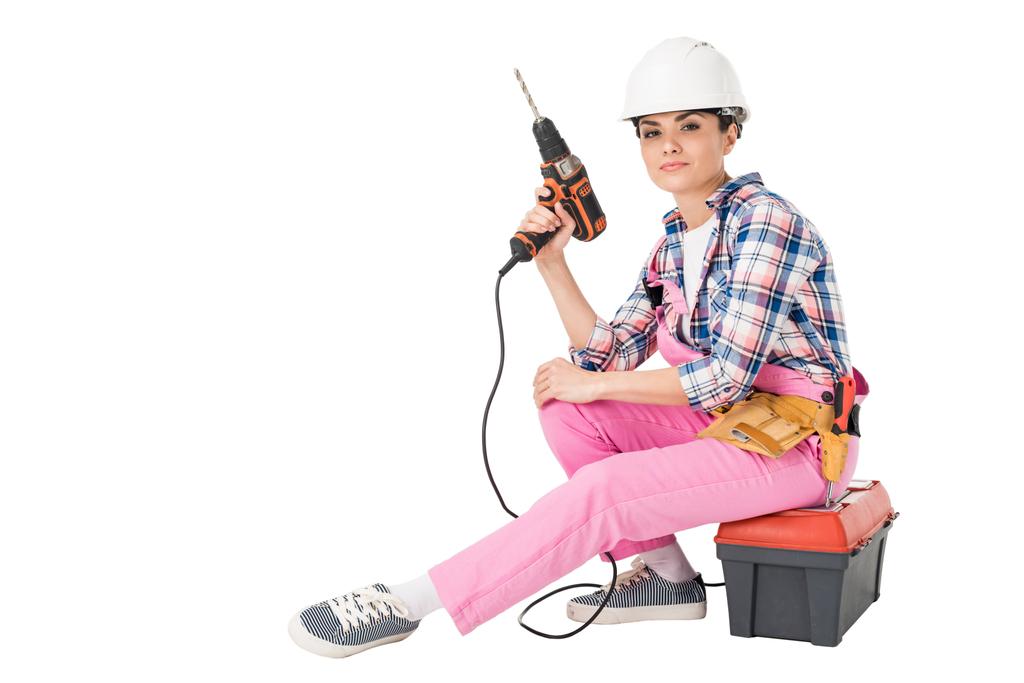 Constructor profesional en overoles y taladro de sujeción de sombrero mientras está sentado en la caja de herramientas aislado en blanco
 - Foto, imagen
