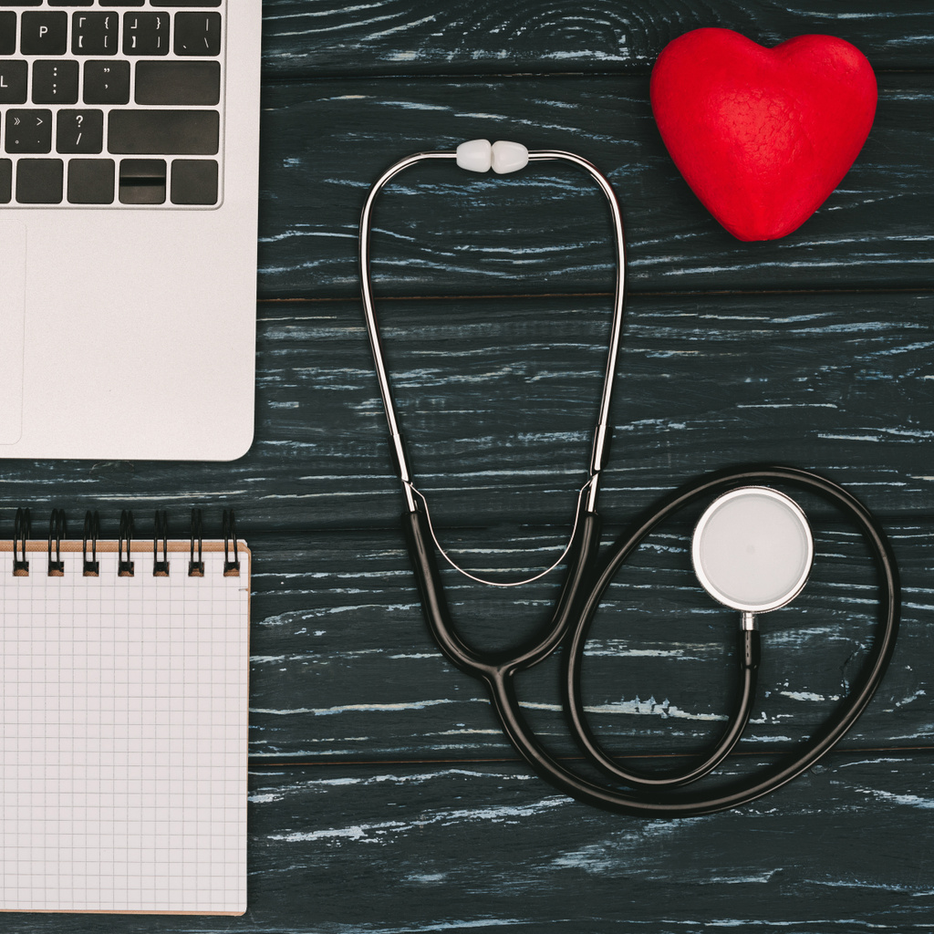 top view of arranged laptop, empty notebook, red heart and stethoscope on dark wooden tabletop - Photo, Image